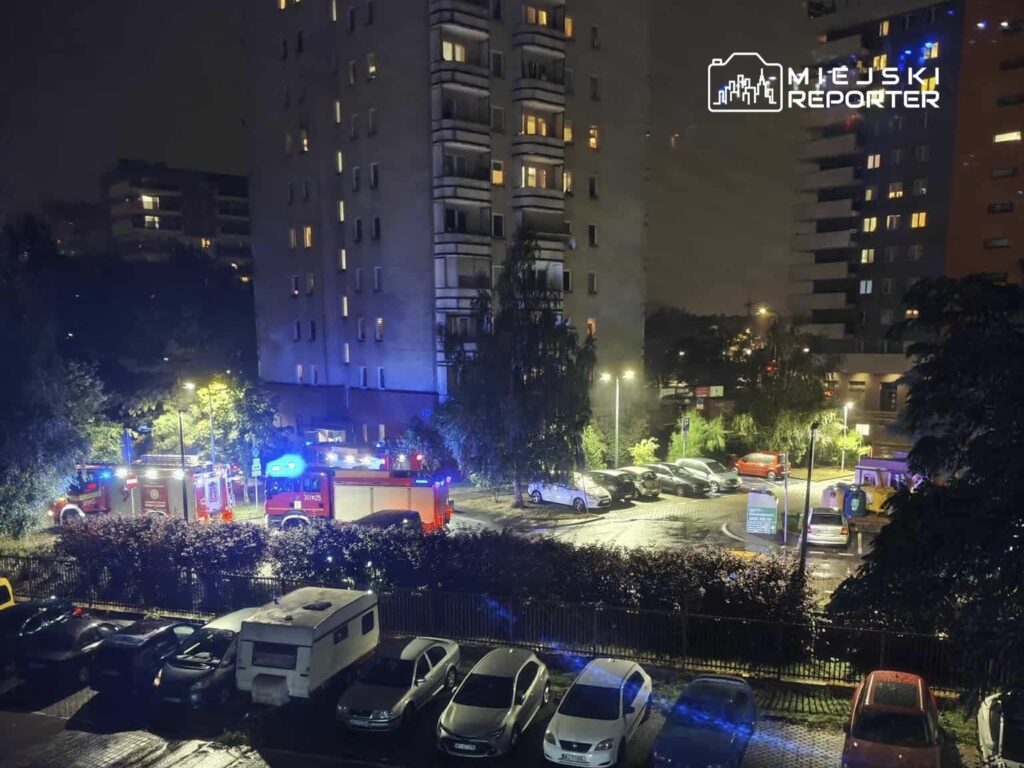 W nocy, w osiedlowym parkingu, straż pożarna z włączonymi syrenami i niebieskimi światłami interweniuje przy budynku mieszkalnym.