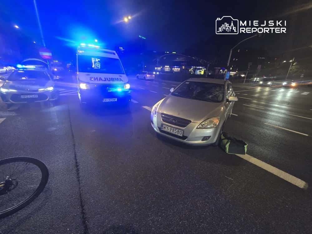 Ambulans z włączonymi sygnałami świetlnymi zatrzymany obok uszkodzonego srebrnego samochodu na ulicy w nocy.