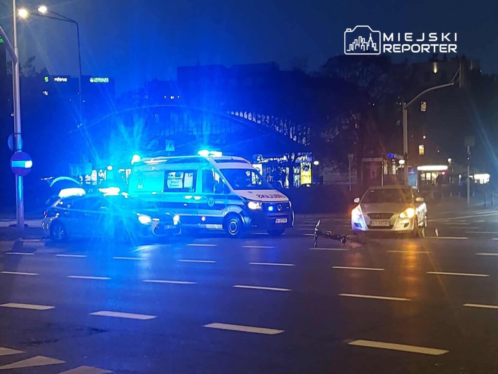 Na skrzyżowaniu ulic stoją samochody policyjne i karetka, w tle widoczne światła nocnego miasta. Na jezdni leży rower.