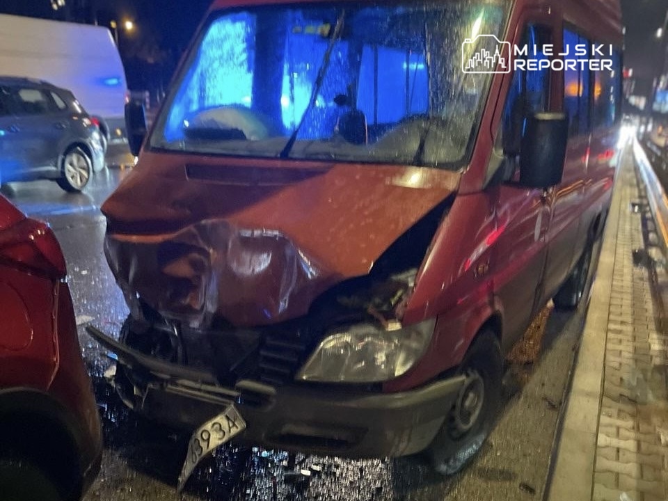 Czerwony samochód dostawczy z uszkodzoną przednią częścią stoi na mokrej ulicy, obok innych pojazdów.