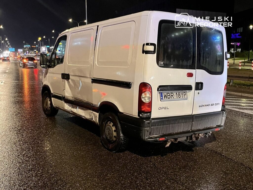 Biały furgon z uszkodzoną boczną częścią stoi na mokrej jezdni w nocy, otoczony innymi pojazdami.