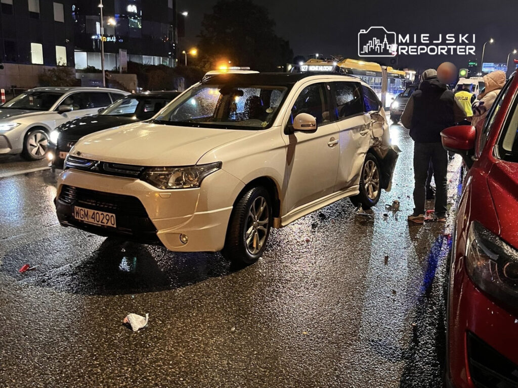 Biały SUV z uszkodzeniami tyłu stoi na mokrej jezdni, obok grupy osób rozmawiających z policją.
