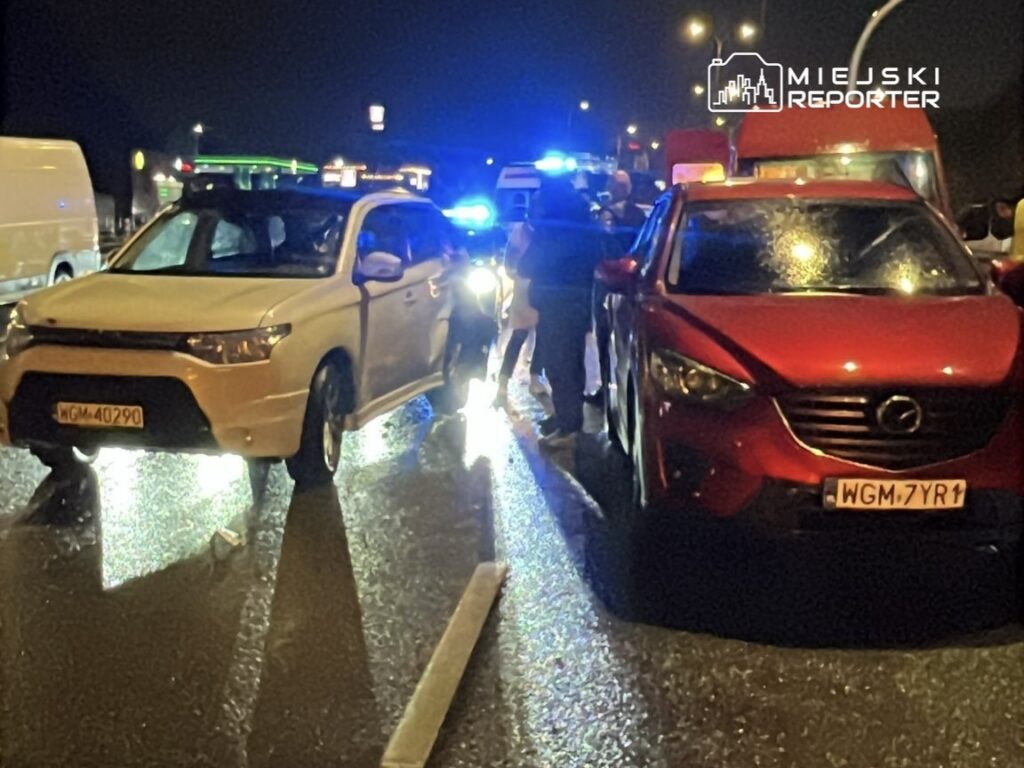 Dwa samochody osobowe zderzone na mokrej drodze w nocy, w tle widać niebieskie światła radiowozów.