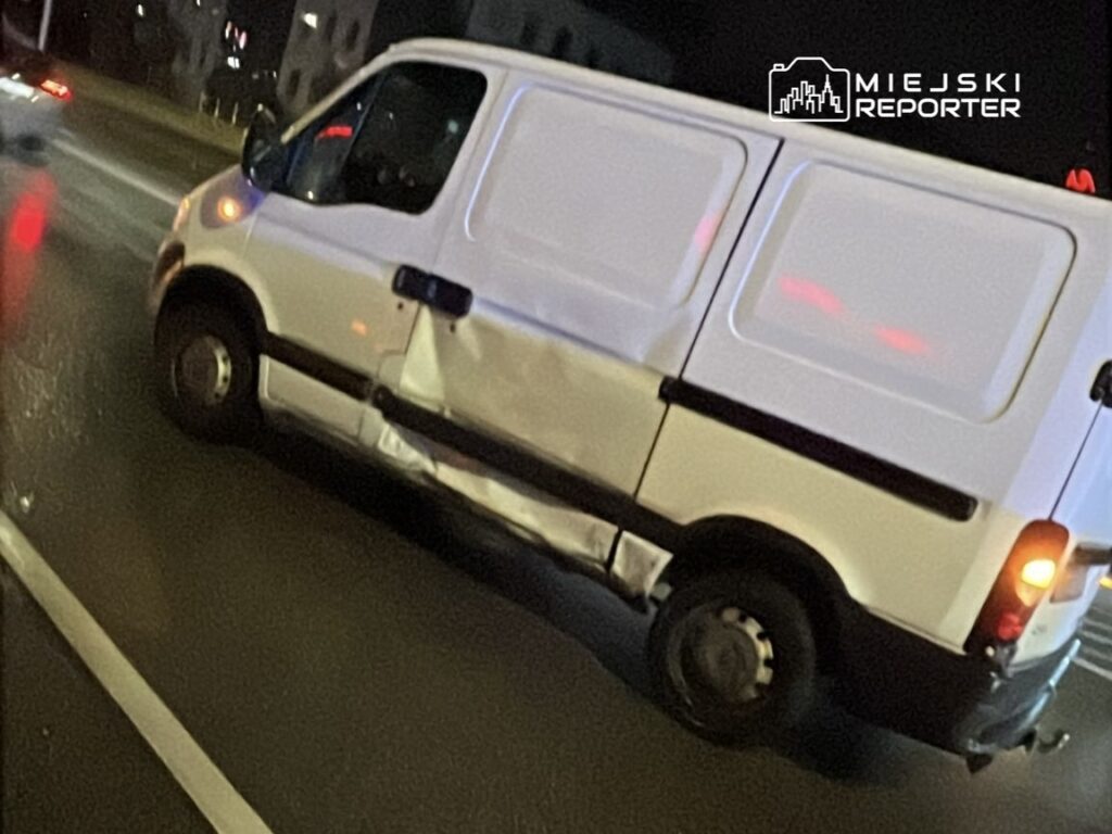 Biały furgon z uszkodzeniami na boku porusza się po drodze w nocy. W tle widać inne pojazdy i oświetlone budynki.