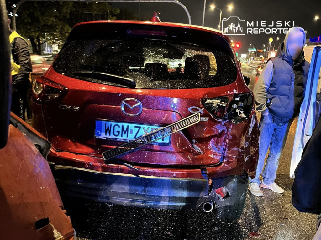 Czerwony samochód osobowy z uszkodzoną tylną częścią stoi na mokrej jezdni, obok mężczyzna w kamizelce odblaskowej rozmawia z innymi osobami.
