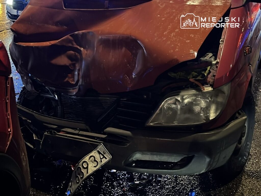 Zniszczony przód czerwonego samochodu po kolizji, z wyraźnymi wgnieceniami i uszkodzeniami, na mokrej nawierzchni drogi.