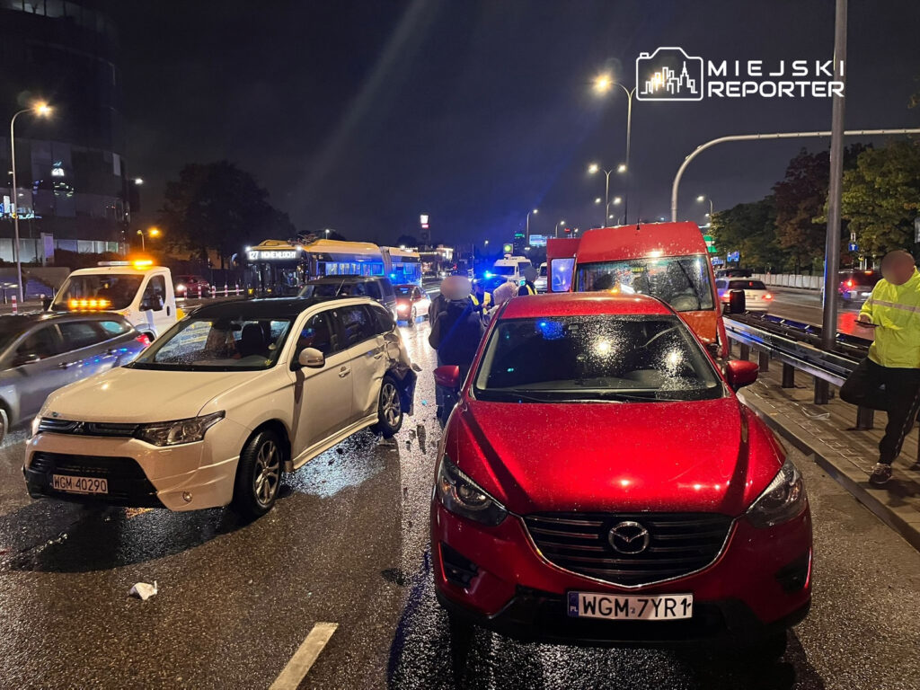 Dwa uszkodzone samochody osobowe na mokrej jezdni, obok nich policjanci i świadkowie zdarzenia w nocy.