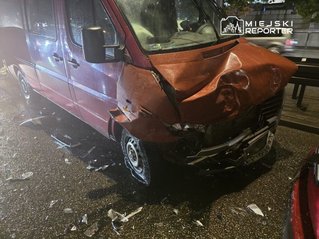Czerwony van z uszkodzoną przednią częścią stoi na ulicy, wokół leżą odłamki szkła i części pojazdów.