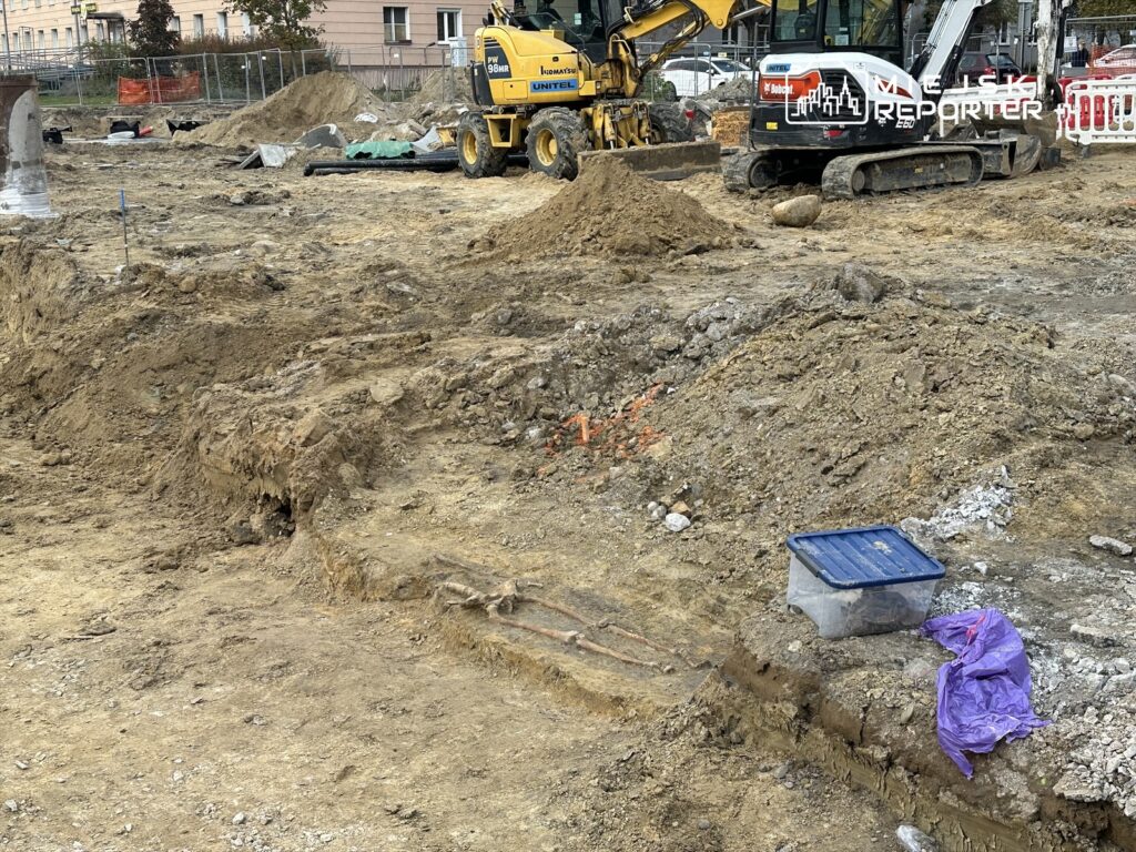 Plac budowy z ciężkim sprzętem, wykopami i porozrzucanym materiałem budowlanym. W tle widoczne budynki mieszkalne.