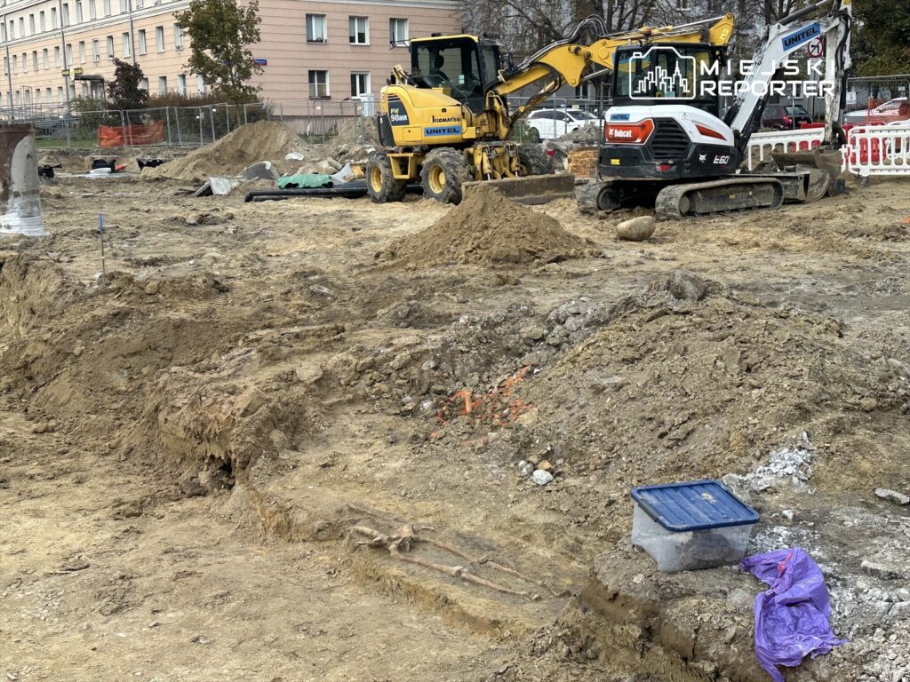 Na placu budowy pracują maszyny budowlane, w tym koparki, w otoczeniu wykopów i materiałów budowlanych.