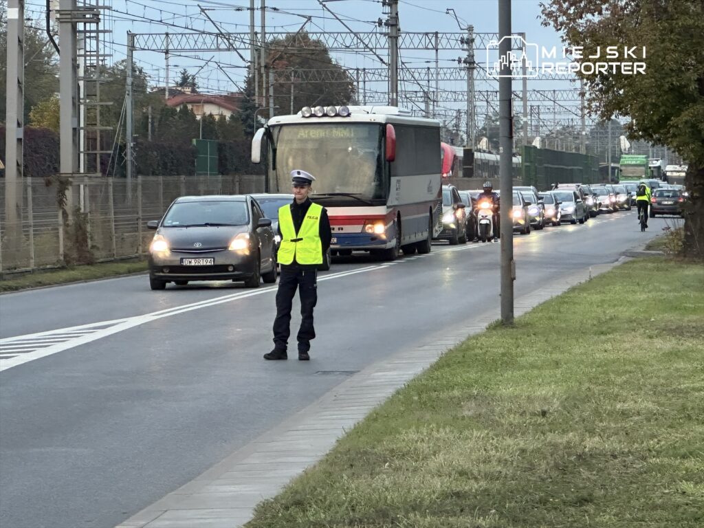 Policjant w żółtej kamizelce kierujący ruchem drogowym na zatłoczonej ulicy, obok przejazdu kolejowego.