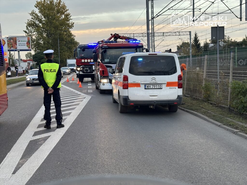 Policjant w kamizelce odblaskowej kieruje ruchem przy wjeździe na drogę, obok pojazd strażacki z włączonymi sygnałami.