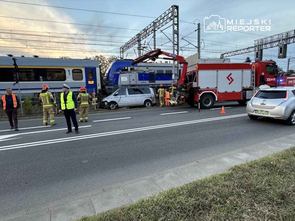 Grupa strażaków i policjant w odblaskowych kamizelkach zabezpiecza miejsce wypadku kolejowego przy drodze.