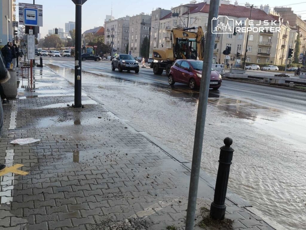 Ruchliwa ulica z samochodami, na której widoczne są kałuże wody i kontener wywożący ziemię, w tle budynki mieszkalne.