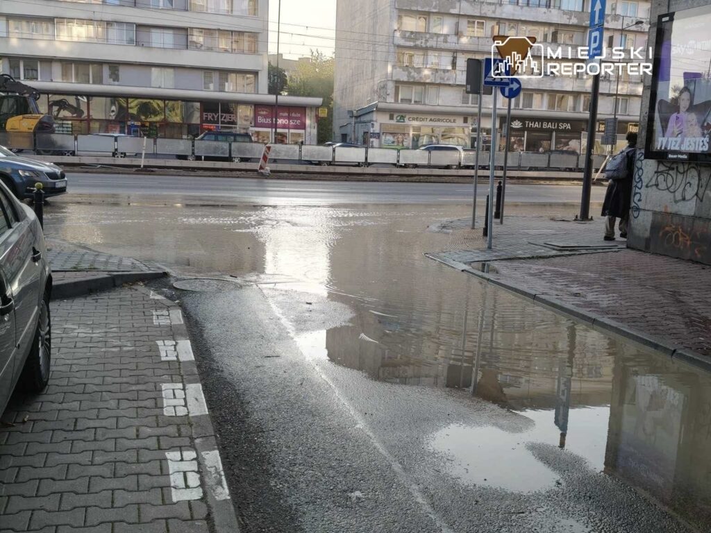 Na ulicy widoczna zalana jezdnia, odbijająca otaczające budynki i znaki drogowe. W tle stoi mężczyzna w koszuli, obserwujący sytuację.