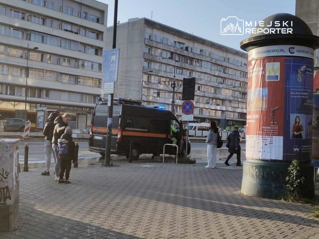 Mężczyzna z dzieckiem na chodniku obok policyjnego vanu z włączonymi sygnałami świetlnymi w miejskim otoczeniu.