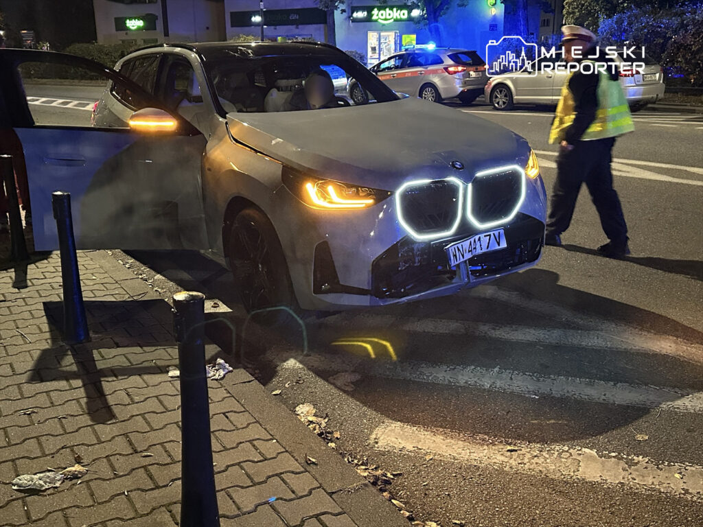 Biały samochód marki BMW z otwartymi drzwiami stoi na chodniku, obok przechodzi policjant w odblaskowej kamizelce.