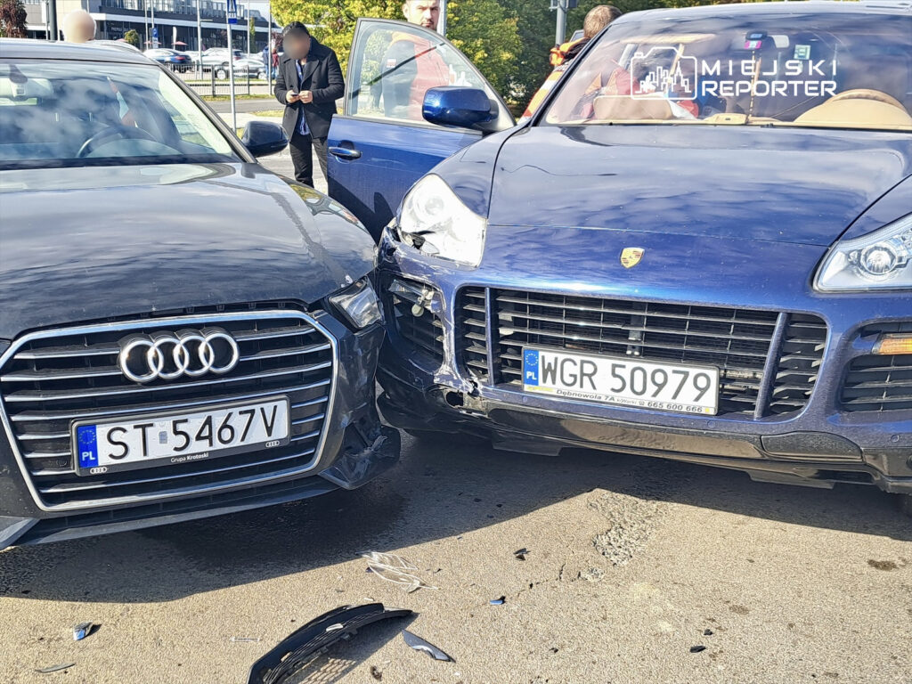 Dwa uszkodzone samochody osobowe stoją obok siebie na parkingu; czarny Audi z uszkodzeniem na przednim lewym boku i niebieski Porsche z uszkodzeniem z przodu. W tle widać ludzi obserwujących sytuację.