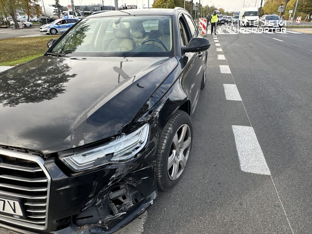 Czarny samochód osobowy z uszkodzoną przednią częścią stoi na drodze, obok widać policję oraz oznakowanie robót drogowych.