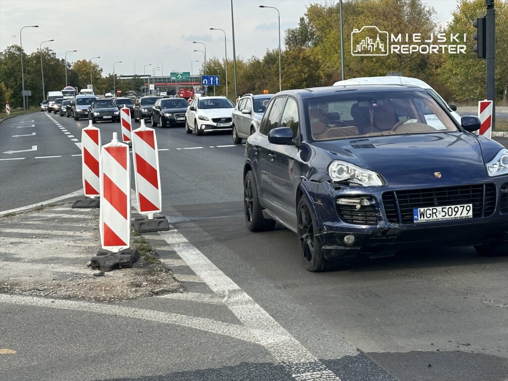 Ruch samochodowy na drodze, z widocznymi barierkami drogowymi i zatrzymanymi pojazdami w tle.