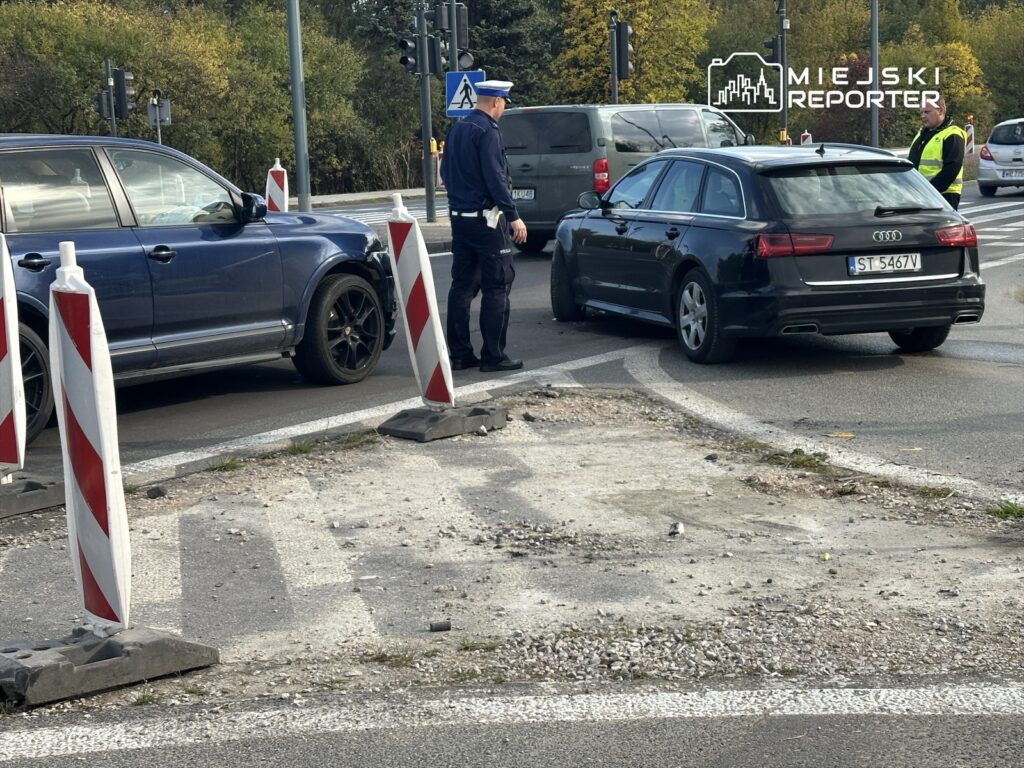 Policjant rozmawia z kierowcą czarnego auta w pobliżu skrzyżowania z sygnalizacją świetlną. W tle widoczne inne pojazdy.