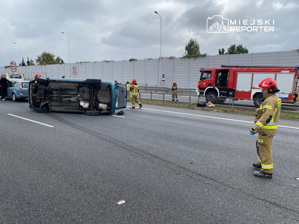 Na drodze leży przewrócony samochód, obok którego pracują strażacy w hełmach i mundurach. W tle widać inne pojazdy oraz woz strażacki.