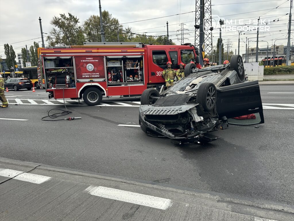 Na jezdni leży przewrócony samochód, obok stoi wóz strażacki z ratownikami w mundurach, a w tle widoczne są inne pojazdy.