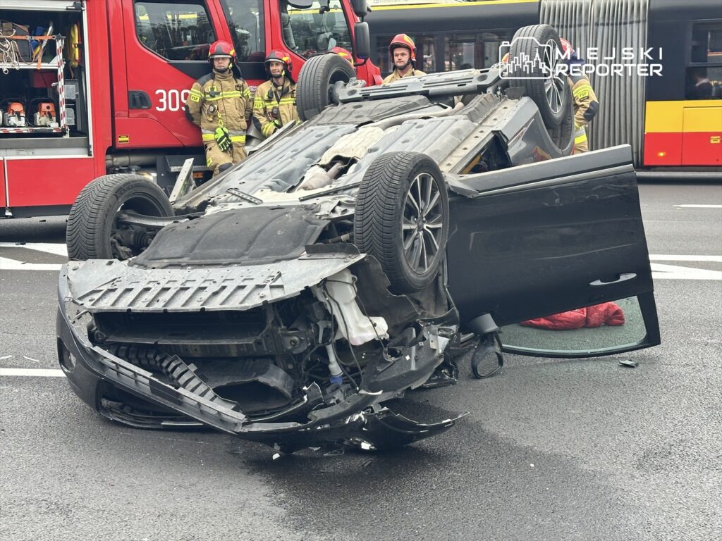Samochód osobowy przewrócony na dach na jezdni, obok stoją strażacy w mundurach, a w tle widoczny autobus.