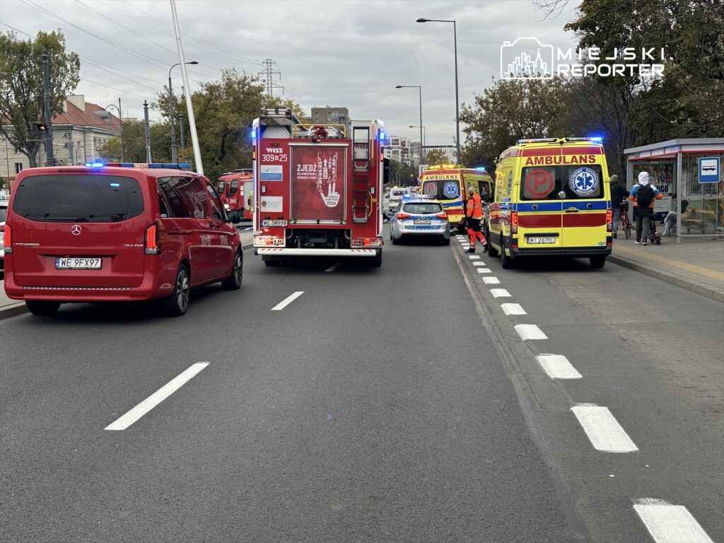 Czerwony van i karetka stoją przy drodze, obok wozu strażackiego, w tle przechodnie czekają na przystanku.