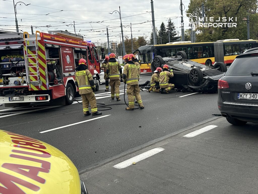 Grupa strażaków w mundurach ratunkowych próbuje przewrócić przewrócone auto na ruchliwej ulicy. W tle widać wóz strażacki i ruch autobusów.