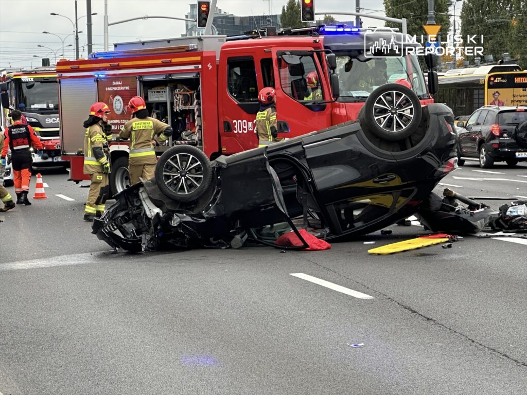 Czarny samochód leży na dachu na jezdni, a strażacy oraz ratownicy medyczni udzielają pomocy poszkodowanym.