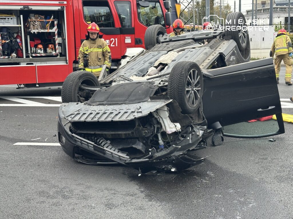 Samochód leżący na dachu na jezdni, obok strażacy w mundurach przygotowujący się do akcji ratunkowej.