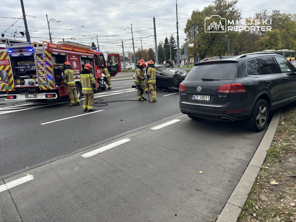 Strażacy pracują nad wypadkiem drogowym, gdzie jeden z pojazdów jest przewrócony na bok.