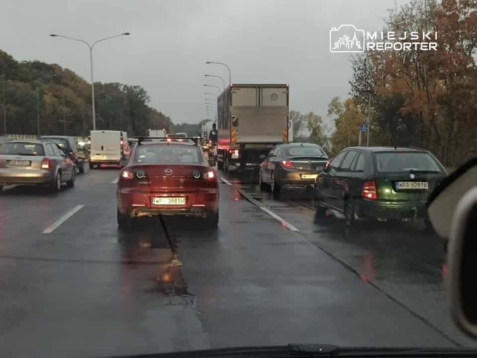 Korkujący się ruch samochodowy na mokrej drodze, z widocznymi pojazdami osobowymi i ciężarówką.