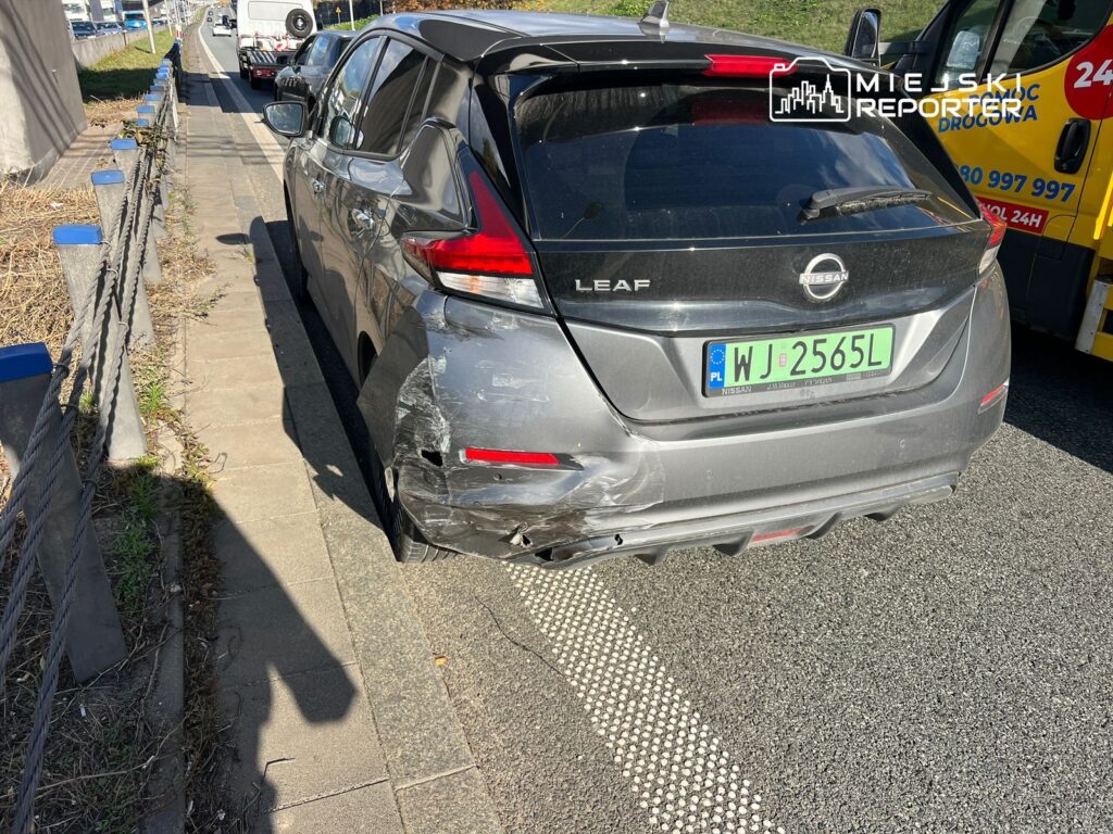 Zniszczony tył samochodu elektrycznego Nissan Leaf zaparkowanego na poboczu drogi, obok przejeżdżających pojazdów.