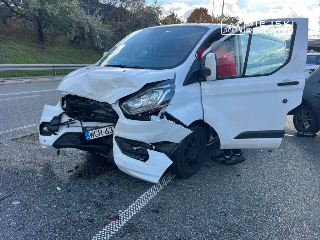 Biały furgon z uszkodzoną przednią częścią stoi na drodze, obok niego widoczny mężczyzna w czerwonej koszulce.