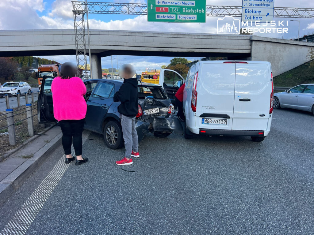 Kobieta w różowym swetrze i mężczyzna w czarnej bluzie rozmawiają obok uszkodzonego samochodu na drodze.