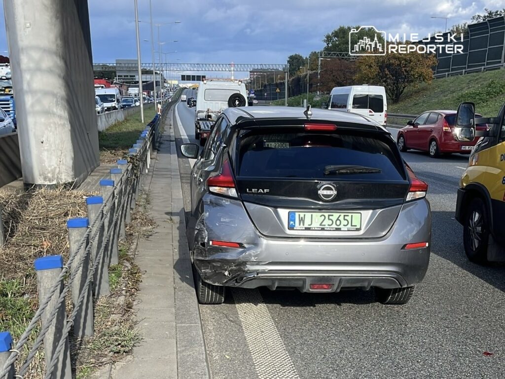 Uszkodzony samochód elektryczny zaparkowany na poboczu ruchliwej drogi, w tle zator drogowy.
