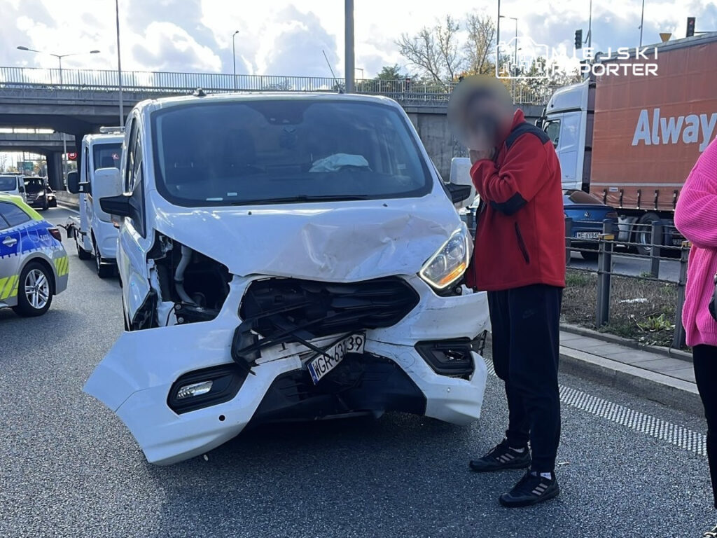 Mężczyzna w czerwonej bluzie stoi obok uszkodzonego furgonu na ruchliwej drodze, a w tle widać inne pojazdy.