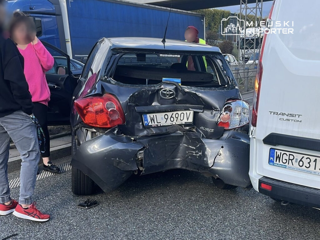 Czarny samochód osobowy z uszkodzoną tylną częścią stoi na poboczu drogi, obok dwóch osób rozmawiających.