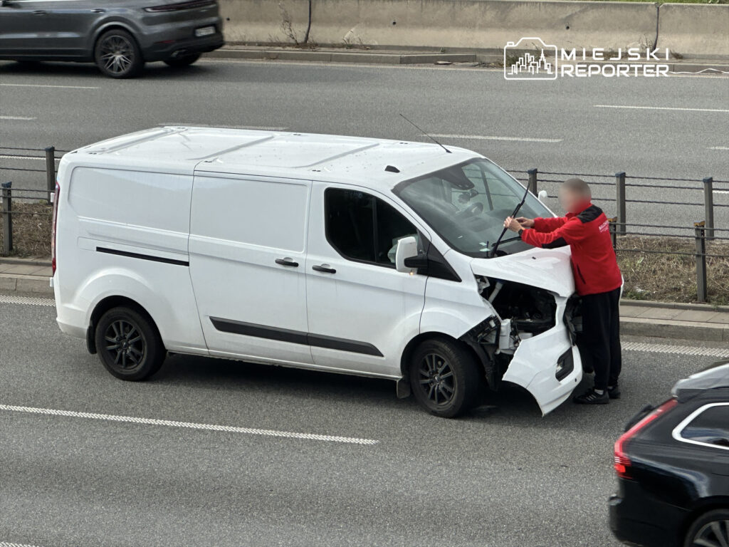 Mężczyzna w czerwonej kurtce naprawia uszkodzony biały samochód dostawczy na poboczu ruchliwej ulicy.