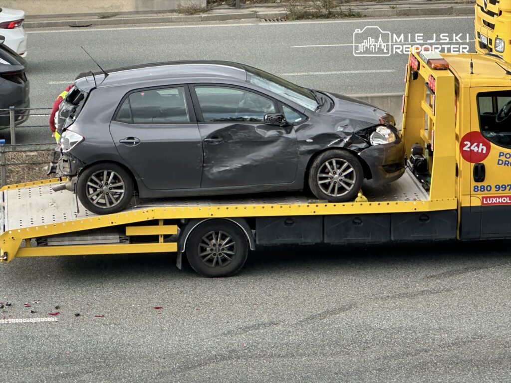 Uszkodzony samochód osobowy na lawecie transportowej na ulicy, obok innych pojazdów.