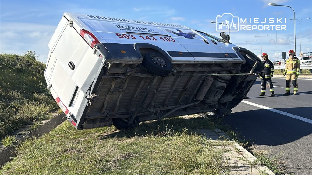 Karetka leży na boku przy drodze, a strażacy pracują nad jej postawieniem.