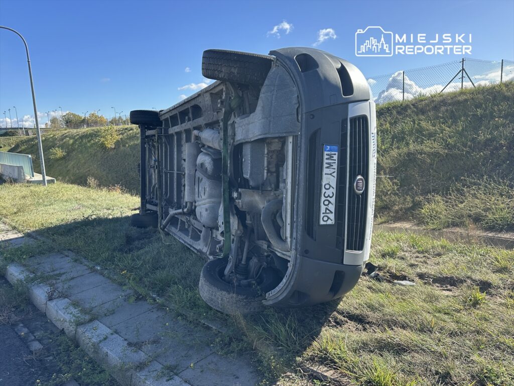 Przewrócony samochód osobowy leży na boku na poboczu drogi, w tle widoczne są drzewa i niebieskie niebo.