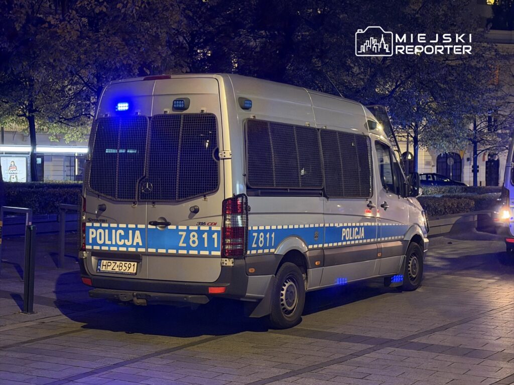 Policyjny samochód dostawczy z niebieskimi światłami zaparkowany w miejskim otoczeniu nocą.