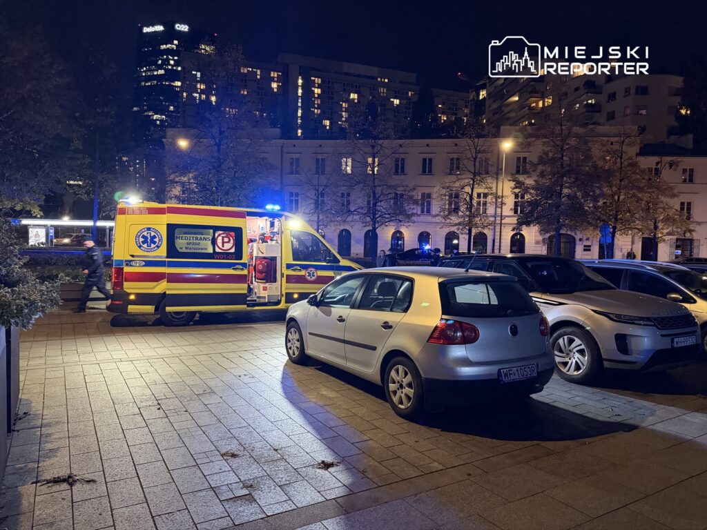 Karetka pogotowia z włączonymi sygnałami świetlnymi zatrzymana na parkingu w nocy, obok zaparkowanych samochodów.