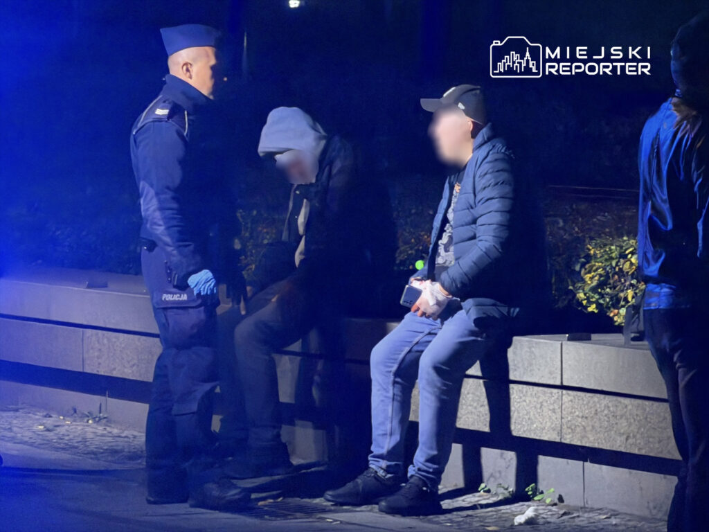 Policjant w mundurze rozmawia z dwoma mężczyznami siedzącymi na ławce w ciemnej okolicy, oświetlonej niebieskim światłem.