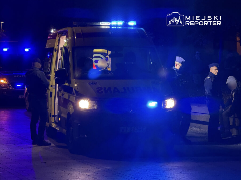 Karetka pogotowia z włączonymi sygnałami świetlnymi na tle policjantów rozmawiających z osobą siedzącą na ławce.