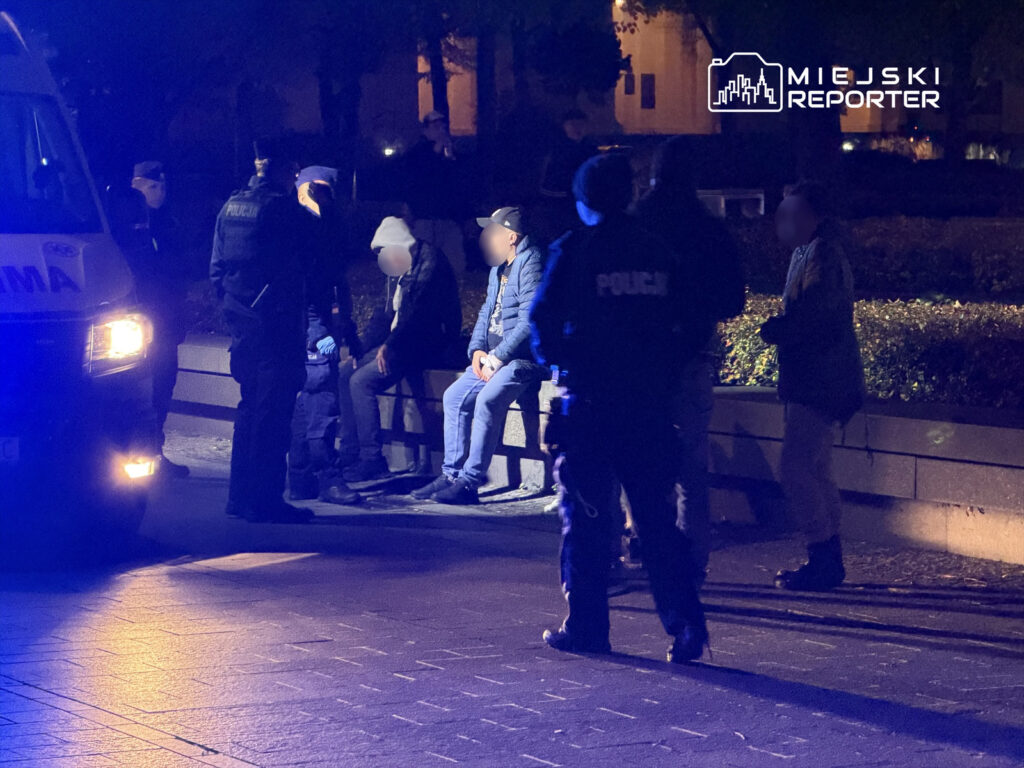 Grupa policjantów rozmawia z siedzącymi mężczyznami na ławce w nocnym, oświetlonym parku.