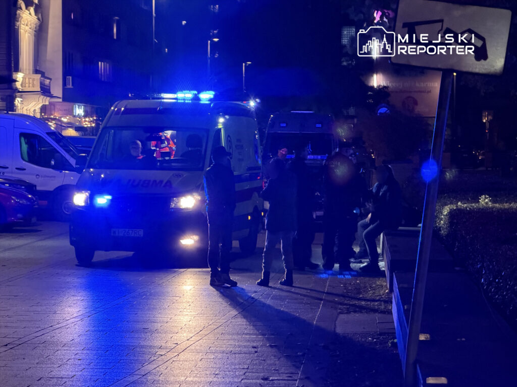 Zespół ratowniczy przy ambulansie z włączonymi sygnałami świetlnymi rozmawia z grupą osób na ulicy nocą.